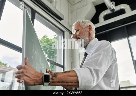 A mature, bearded teacher in a white shirt writes equations on a whiteboard, teaching online. Stock Photo