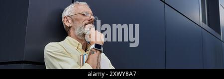 A mature man with a beard and glasses leans against a modern building, lost in thought. Stock Photo