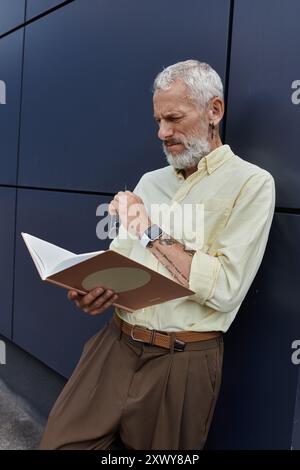 A bearded, middle-aged man in a yellow shirt reads a book while standing against a modern building. Stock Photo