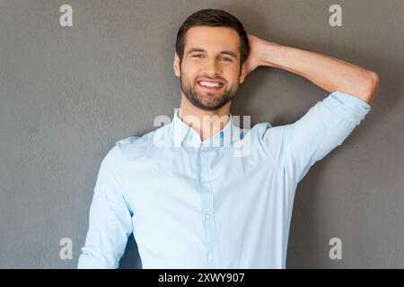 Feeling satisfied and relaxed. Handsome young man in shirt touching his head and smiling at camera while standing against grey background Stock Photo