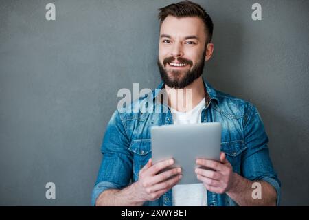 Smart technology makes everything easier. Happy young man holding digital tablet and smiling at camera while standing against grey background Stock Photo