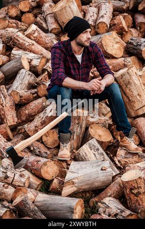 Taking smoking pause. Full length of pensive young forester smoking and looking away while sitting on logs Stock Photo
