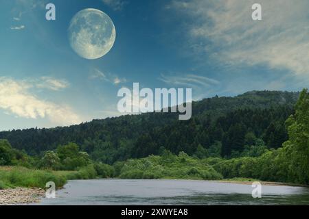 Highly detailed bright full moon (supermoon) in the twilight or night cloudy blue sky. Mountains and river panoramic landscape. Dramatic and picturesq Stock Photo