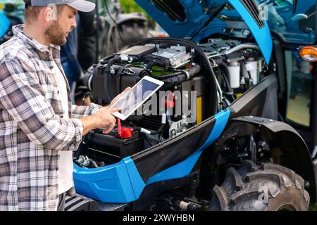 Man agricultural machinery mechanic diagnoses tractor engine operation using digital tablet. Stock Photo