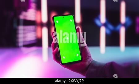 Anonymous Man Holding a Smartphone with Mock Up Green Screen Chromakey Display in a Futuristic Room with Neon Lights. Photo Template with Placeholder for Tech Advertising and Entertainment Content Stock Photo