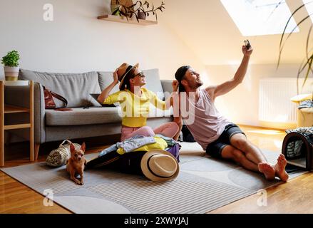 Young joyful couple with his small dog packing their travel bag and taking a selfie using phone. Stock Photo