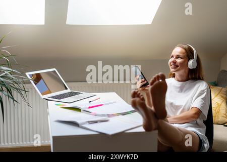 Young woman student wearing white t-shirt woman studying at a desk at home. Listen to educational podcast. Stock Photo