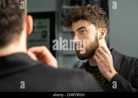 Handsome young man with beard looking in mirror, touching his face. Concept of male grooming, self-care routine, and modern masculinity. Stylish hairc Stock Photo