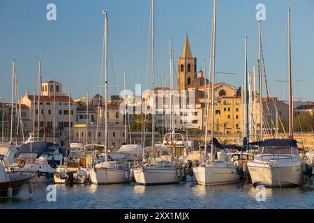 Yachts moored in marina beneath the Old Town walls, sunrise, bell tower of the cathedral prominent on skyline, Alghero, Sassari, Sardinia Stock Photo