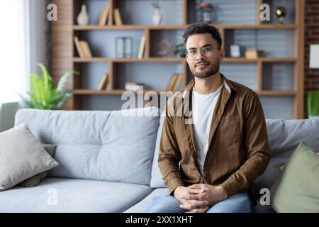 Portrait of young serious Indian man sitting on sofa at home in casual clothes and looking at camera. Stock Photo