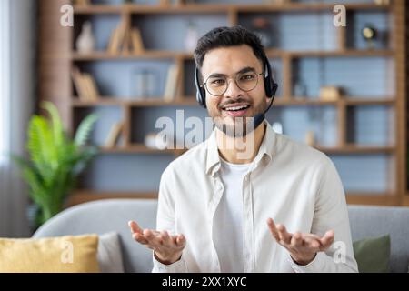 Portrait of a young hispanic man in a headset and glasses sitting on the couch at home and talking to the camera, while gesturing with his hands. Stock Photo