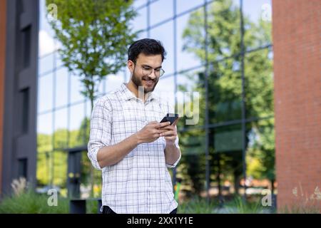 Smiling young man using phone outside modern office building with glass facade. Standing next to tree and brick wall on sunny day, enjoying conversation or browsing internet. Stock Photo