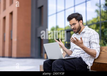 Young man shows frustration while working on laptop outdoors. Urban setting with modern office buildings in background. Difficulties with technology, work stress, and problem solving portrayed Stock Photo