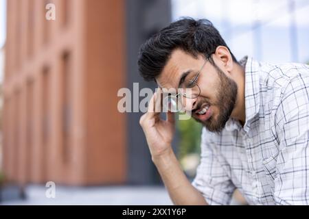 Young man wearing glasses experiencing stress or tiredness while outdoors. Man has hand on his head showing frustration or headache in front of a modern building. Stock Photo