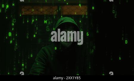 A hooded hacker in a dark hoodie sits in a dimly lit room, surrounded by glowing green code cascading down the screen. The secretive and shadowy world Stock Photo