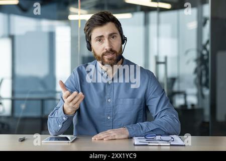 Portrait of a young man in a headset sitting in the office at the table and talking to the camera, explaining and gesturing with his hands. Stock Photo