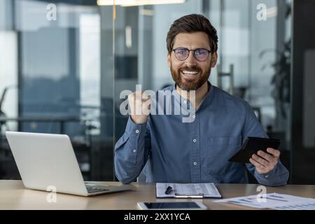 Portrait of a young man in glasses sitting in the office at the desk and enjoying financial success, holding a calculator, showing a yes gesture with his hand, looking at the camera. Stock Photo