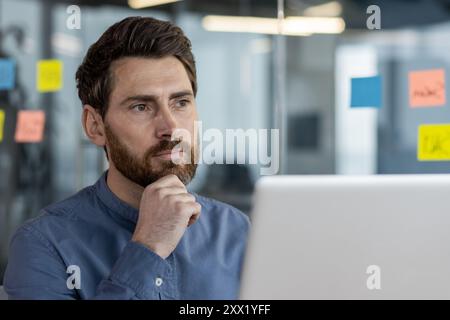 Close-up photo of a thoughtful and serious young man sitting in the office at the workplace in front of the laptop, thoughtfully looking ahead, holding his head with his hand. Stock Photo