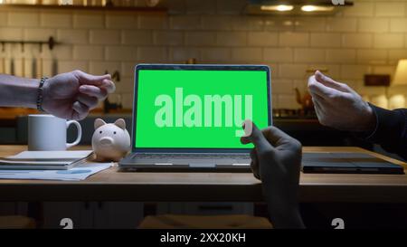 Laptop computer with mock up green screen chromakey display standing on the kitchen table. Anonymous hands doing money gesture, background modern kitc Stock Photo