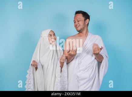 Smiling young Asian Muslim couple wearing ihram and prayer gown with praying hand gesture over isolated by blue background. Hajj concept. Stock Photo