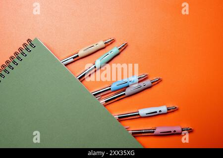 A green spiral notebook with a row of pens on top of it. The pens are of different colors and sizes, and they are lined up neatly Stock Photo