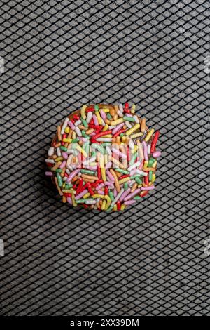 A colorful mini donut covered in sprinkles sits on a metal surface. The sprinkles are in various colors and are scattered all over the mini donut. Con Stock Photo