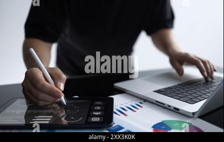 A professional works on a tablet displaying infographics while also using a laptop, surrounded by financial charts and documents. The image illustrate Stock Photo