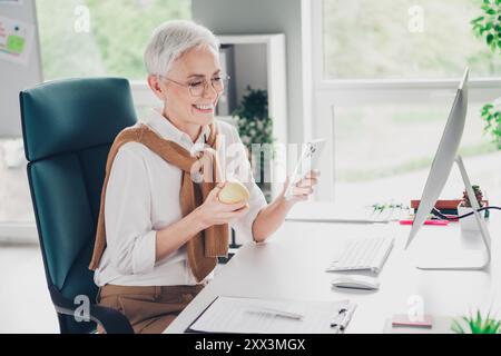 Photo of nice boss mature woman eat apple lunch device break dressed formalwear comfortable light startup office loft room interior Stock Photo