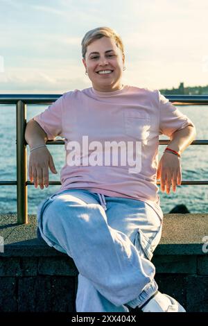 Cheerful plus-size woman with short hair and nose piercing sits on waterfront railing, enjoying sunset. Body positivity, self-acceptance, and diversit Stock Photo