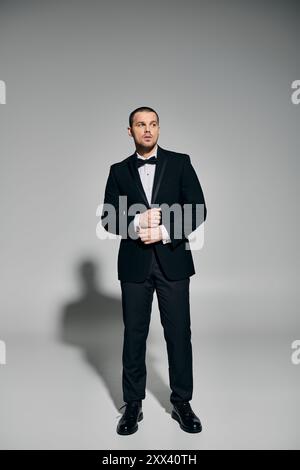 A man in a black tuxedo stands against a grey backdrop, looking up and adjusting his jacket. Stock Photo