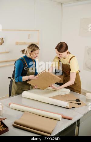 Two young creative female workers of design studio or craft shop packing and wrapping parcel into paper by workplace Stock Photo
