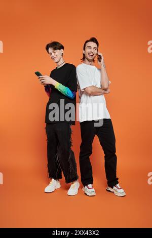 Two stylishly dressed queer friends stand against an orange backdrop, one using a phone while the other chats on one. Stock Photo