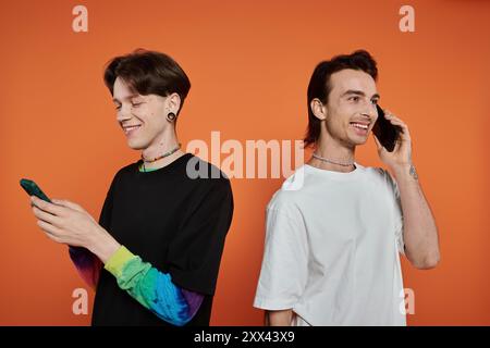 Two stylishly dressed queer friends against a vibrant orange background; one texting while the other is on a call. Stock Photo