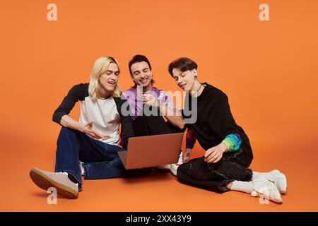 Three young friends in colorful attire sit on an orange background, looking at a laptop. Stock Photo
