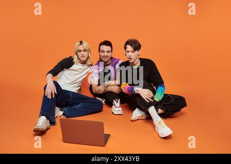Three young queer friends in vibrant attire are seated on an orange backdrop, watching something on a laptop. Stock Photo