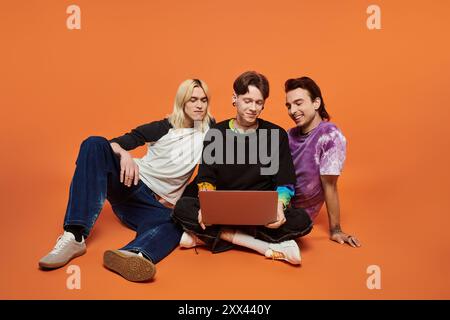 Three young queer friends in stylish, colorful clothing sit on a bright orange background, one using a laptop. Stock Photo
