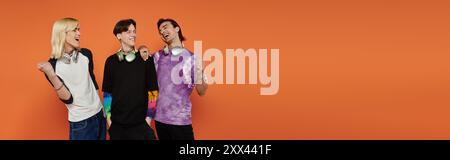 Three friends in stylish attire stand against an orange background and laugh. Stock Photo