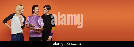 Three young friends in stylish attire stand against an orange background. They are engaged in a conversation. Stock Photo