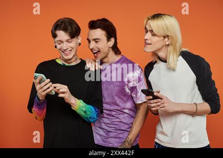 Three young friends in stylish, vibrant attire laugh together against an orange background. Stock Photo