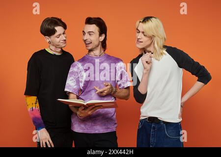 Three young friends in stylish, colorful attire stand against an orange background, laughing and sharing a book. Stock Photo