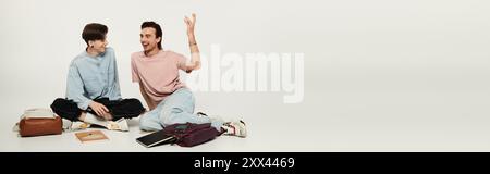Two students in casual attire sit on the floor against a white backdrop, laughing and chatting, displaying their bond. Stock Photo