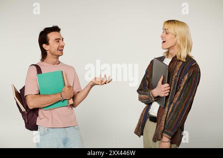 Two young queer students stand together, one holding a notebook and the other a laptop, sharing a laugh against a grey background. Stock Photo