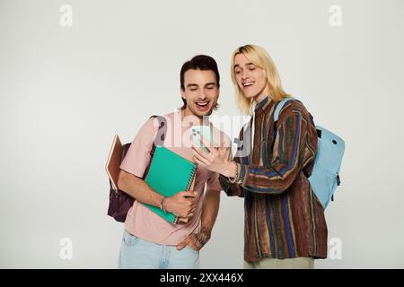 Two young students, dressed casually, stand against a grey background and share a laugh while looking at a phone screen. Stock Photo