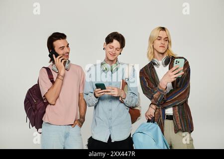 Three young queer students stand together in casual attire, using their phones and laughing. Stock Photo