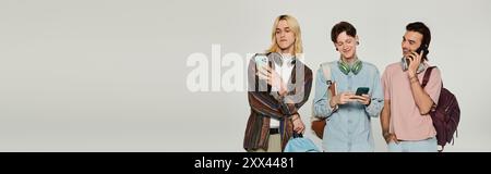 Three young queer students stand together, using their phones, in front of a grey backdrop. Stock Photo