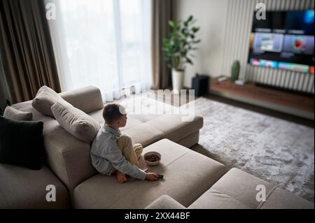 Lazy morning for little boy. Cute kid eating cereals and watching favorite TV show. Joyful child relaxing on couch in living room and watching television. Stock Photo