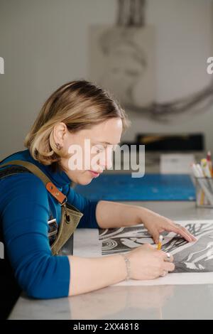 Side view of young female artisan in blue pullover sitting by desk in workshop and making sketch of artwork for printing Stock Photo