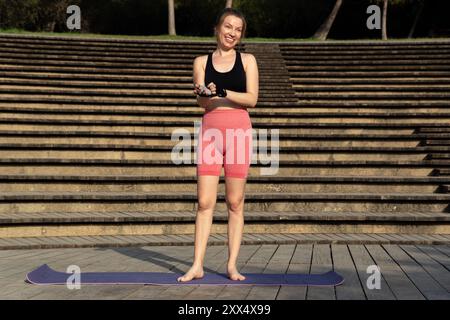 A vibrant young redhead woman, dressed in sporty attire and gloves, stands on a wooden deck under the sun, smiling as she prepares for her outdoor yog Stock Photo