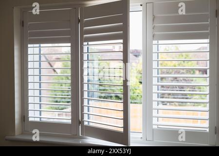 Three frames of  white modern wooden shutters across a window. One section is open. The shutter slats are swivelled in different positions. Stock Photo