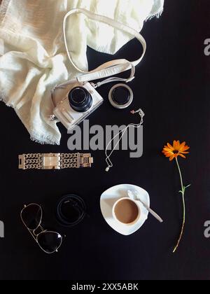 Vertical Flat Lay of Woman’s Elegant Accessories: Camera, Scarf, Coffee, Watch, Sunglasses, Pendant, Lens, Belt, and Orange Flower on Black Table Stock Photo